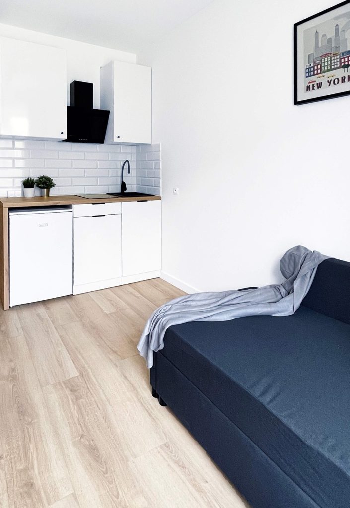 Nowoczesna kuchnia w białej zabudowie z drewnianym blatem i czarnym okapem. Obok znajduje się ciemnoszara sofa z narzuconym kocem. Jasne wnętrze z minimalistycznym designem.