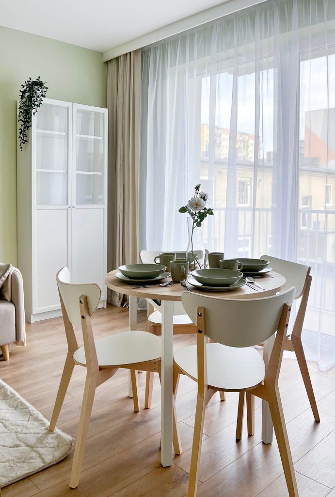 Salon z jadalnią – oliwkowa ściana, okrągły stół, białe krzesła z drewnianymi nogami, jasna sofa i duże okno balkonowe z wyjściem na balkon - mieszkanie 1-pokojowe na wynajem w Łodzi, dzielnica Bałuty