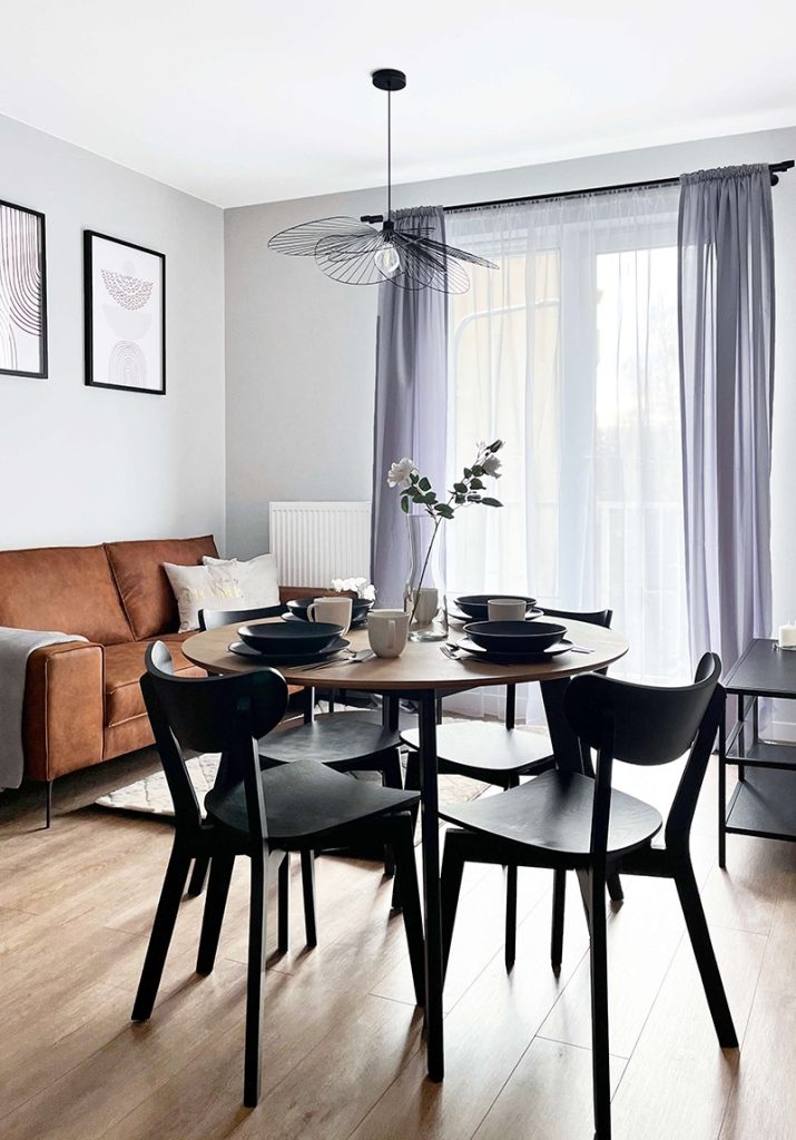 Jasny salon z jadalnią – okrągły stół, czarne krzesła, brązowa sofa i wyjście na balkon z fioletowymi zasłonami - mieszkanie 3-pokojowe na wynajem w Łodzi, dzielnica Bałuty