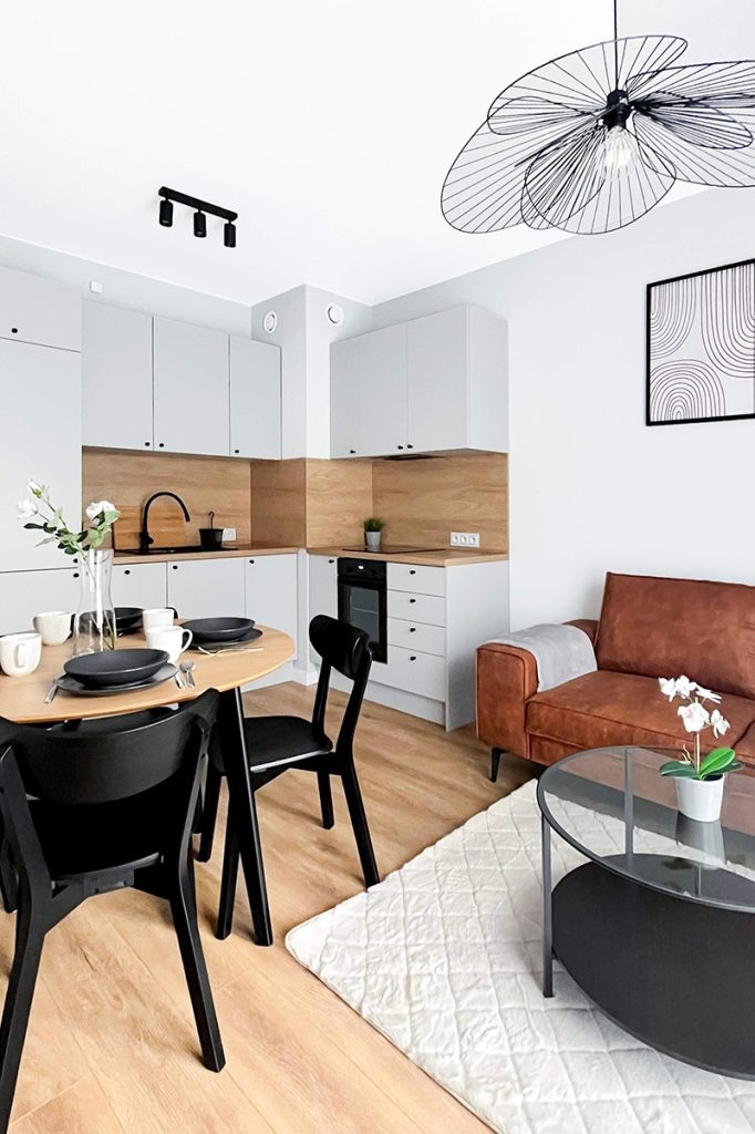 Salon z aneksem kuchennym – okrągły stół jadalniany, czarne krzesła, brązowa sofa i zabudowa kuchenna w odcieniu szarości z drewnianym wykończeniem. 3 pokojowe mieszkanie na wynajem - Łódź