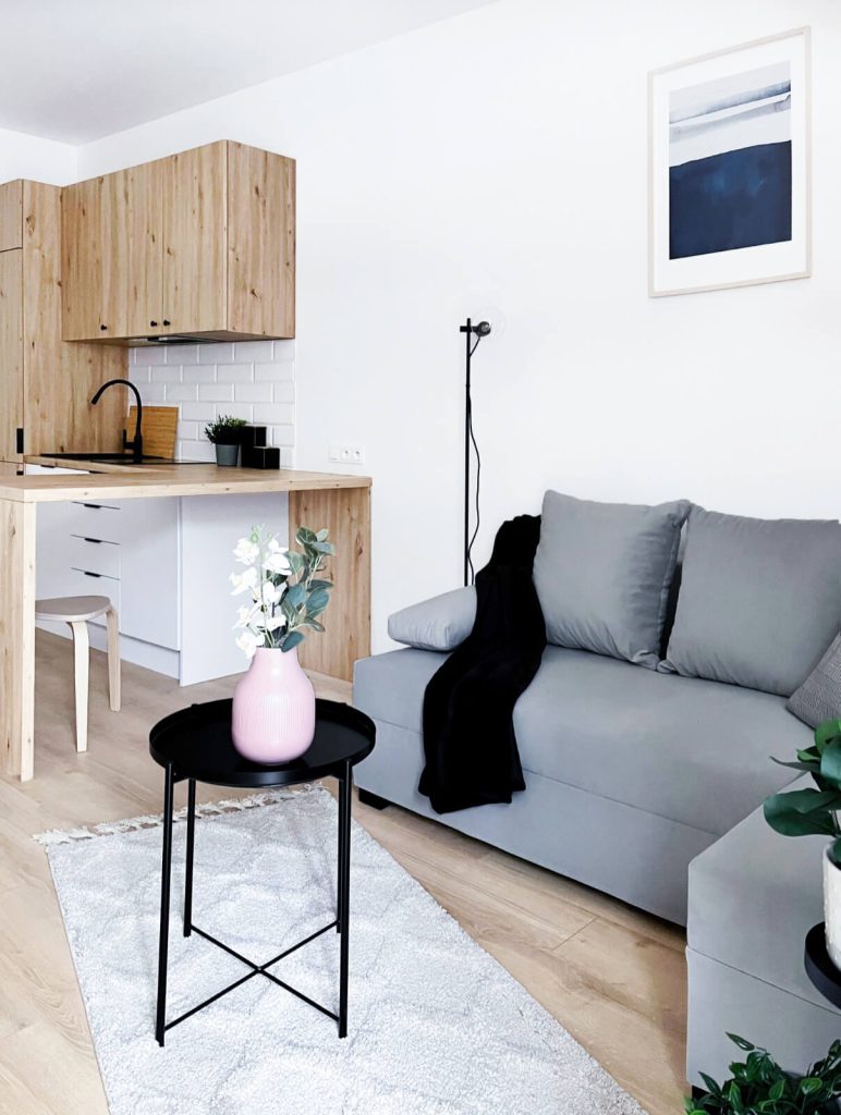 Nowoczesny salon z szarą sofą, stolikiem kawowym i jasną kuchnią z drewnianymi frontami i białą cegiełką na ścianie - mieszkanie na wynajem Łódź Śródmieście