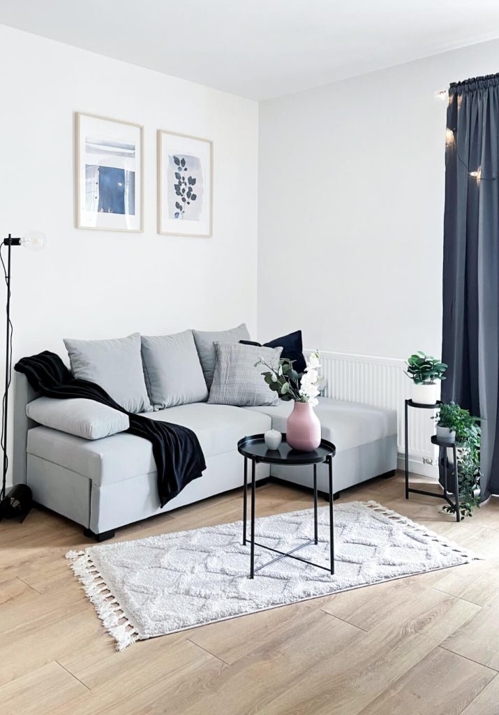 Część dzienna w kawalerce na wynajem w Łodzi z szarą sofą i czarnym stolikiem kawowym. Jasne wnętrze z drewnianą podłogą i granatowymi zasłonami