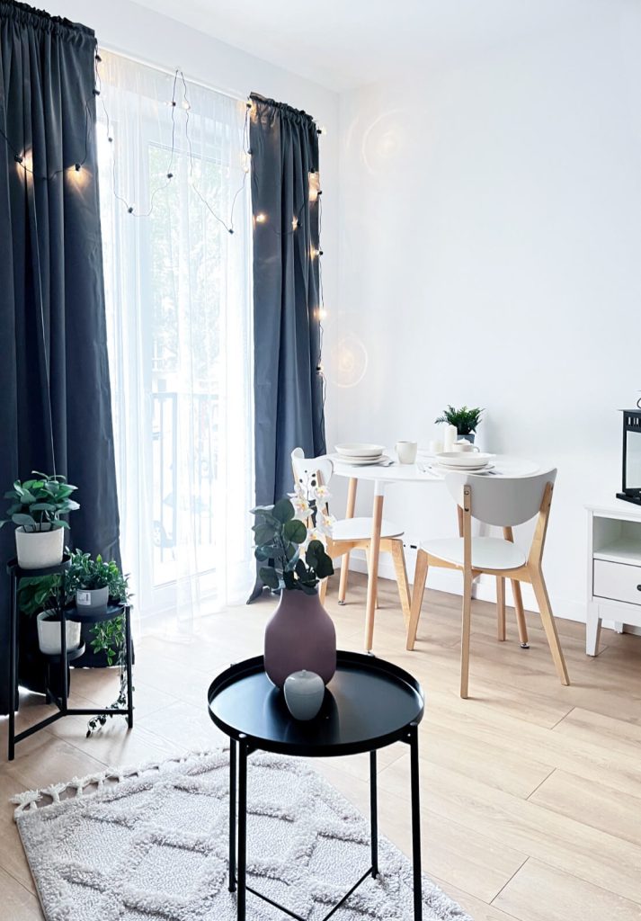 Przytulna kącik jadalny z białym stołem, krzesłami z jasnego drewna i dużym oknem balkonowym i ciemnymi zasłonami - kawalerka na wynajem Łódź Pomorska 107a