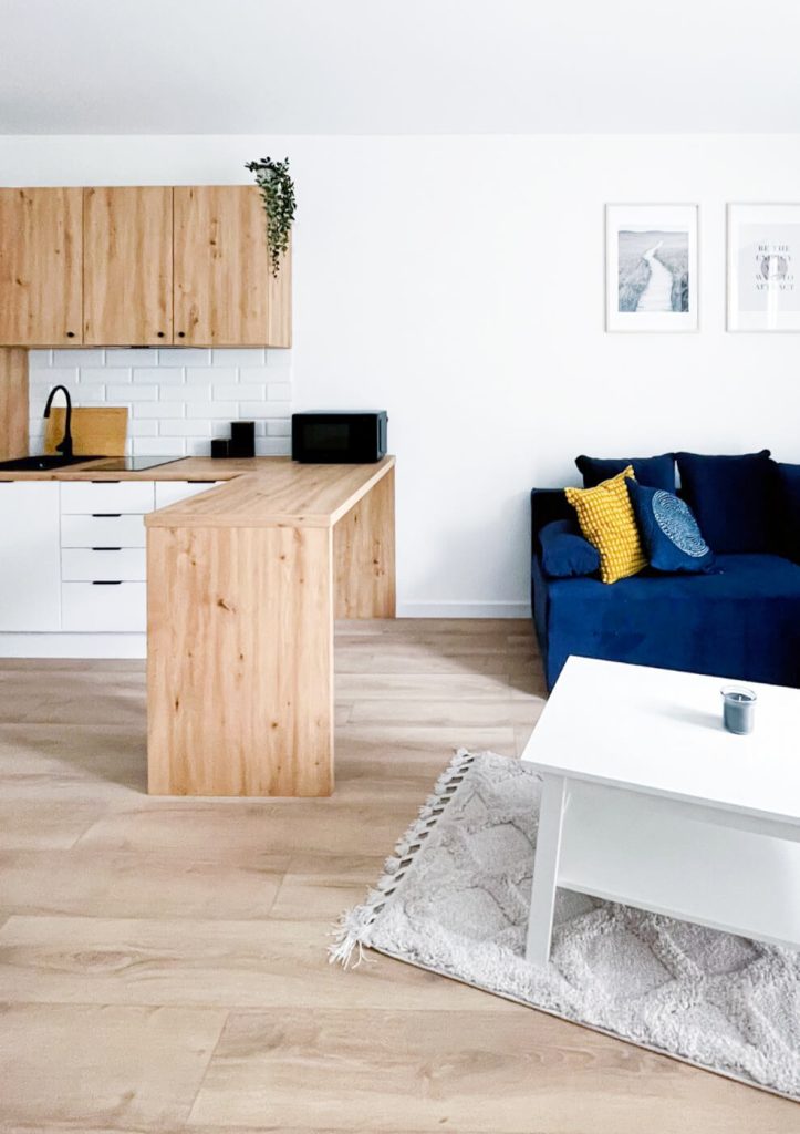 Nowoczesny salon z granatową sofą, białym stolikiem kawowym i jasną kuchnią z drewnianymi frontami i białą cegiełką na ścianie - mieszkanie na wynajem Łódź Śródmieście