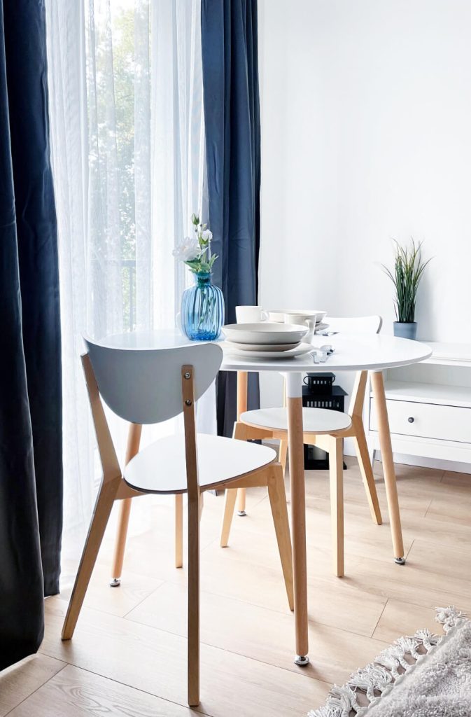 Minimalistyczny stół jadalniany z dwoma białymi krzesłami ustawiony przy dużym oknie z ciemnymi zasłonami - kawalerka na wynajem Łódź Pomorska 107a