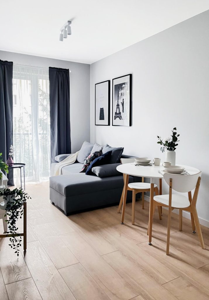 Widok na salon z ciemną sofą z ozdobnymi poduszkami, okrągły biały stół, z krzesłami z jasnego drewna i dużym oknem balkonowym i ciemnymi zasłonami - mieszkanie 2-pokojowe na wynajem Łódź Pomorska 107a