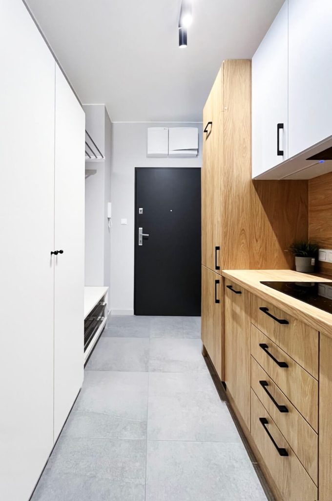 Widok na przedpokój mieszkania z czarnymi drzwiami wejściowymi, jasnoszarą podłogą i nowoczesną zabudową kuchenną w kolorze drewna i bieli z czarnymi uchwytami - mieszkanie na wynajem w Łodzi, Śródmieście