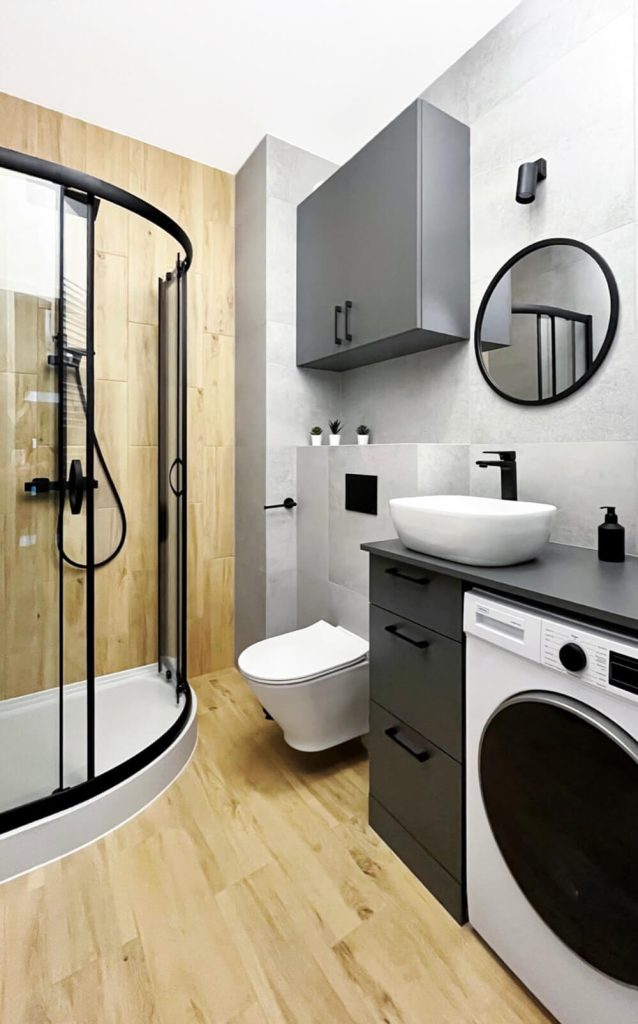 Łazienka z prysznicem, podwieszana toaleta, pralką, czarną armaturą i drewnopodobnymi akcentami – połączenie szarości i drewna - mieszkanie na wynajem Łódź, Śródmieście