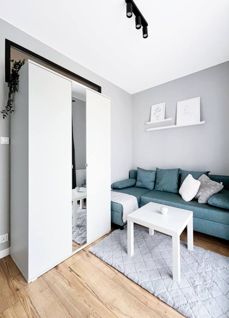 Pokój z narożną sofą w kolorze niebieskim, białym stolikiem kawowym i dekoracyjnymi grafikami nad kanapą oraz białą szafą - mieszkanie na wynajem Łódź Śródmieście