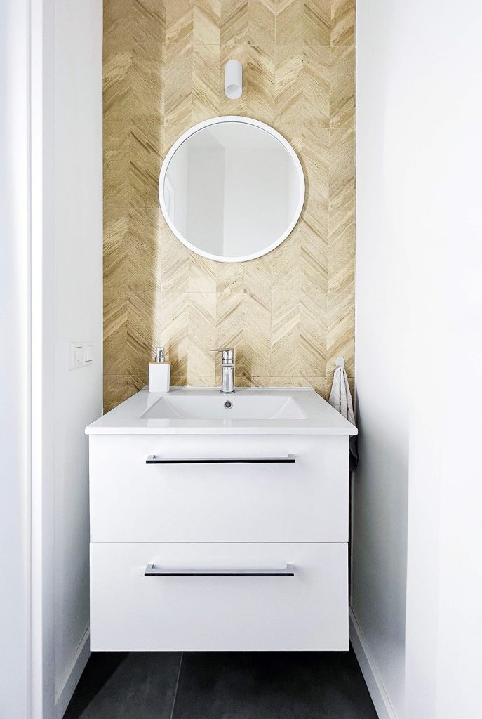 Nowoczesna biała umywalka z szufladami, okrągłym lustrem i lampką ścienną, na tle ściany z drewnopodobnych płytek w jodełkę - pokoje na wynajem w mieszkaniu 3 pokojowym w Łodzi (cześć wspólna)