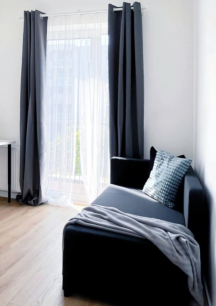 Jasny pokój na wynajem z dużym oknem, ciemną sofą z poduszką i kocem, ozdobiony firankami i ciemnymi zasłonami - pokój na wynajem w mieszkaniu 3 pokojowym w Łodzi Śródmieście