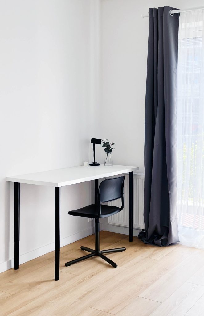 Jasny pokój, białe biurko z ciemnymi metalowymi nogami, czarna lampka na biurku, czarne krzesło, ciemna zasłona przy dużym oknie - pokój na wynajem w mieszkaniu 3 pokojowym w Łodzi Śródmieście